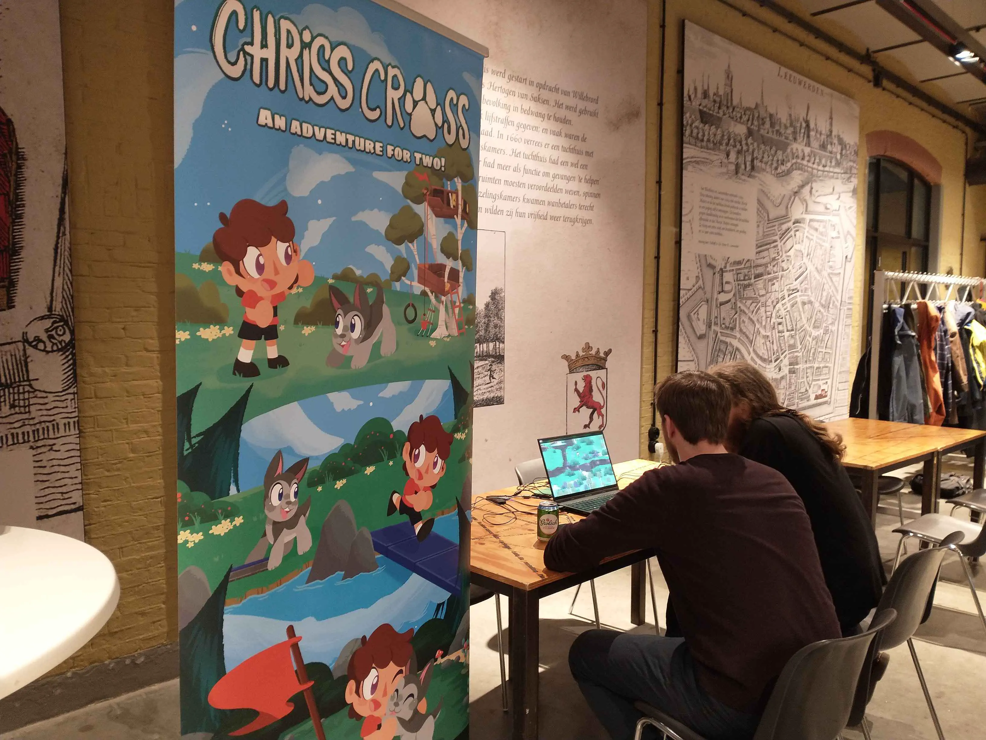 A photo taken during the Open Dev Mix, a banner of Chriss Cross is visible, two people are playing a game behind a laptop