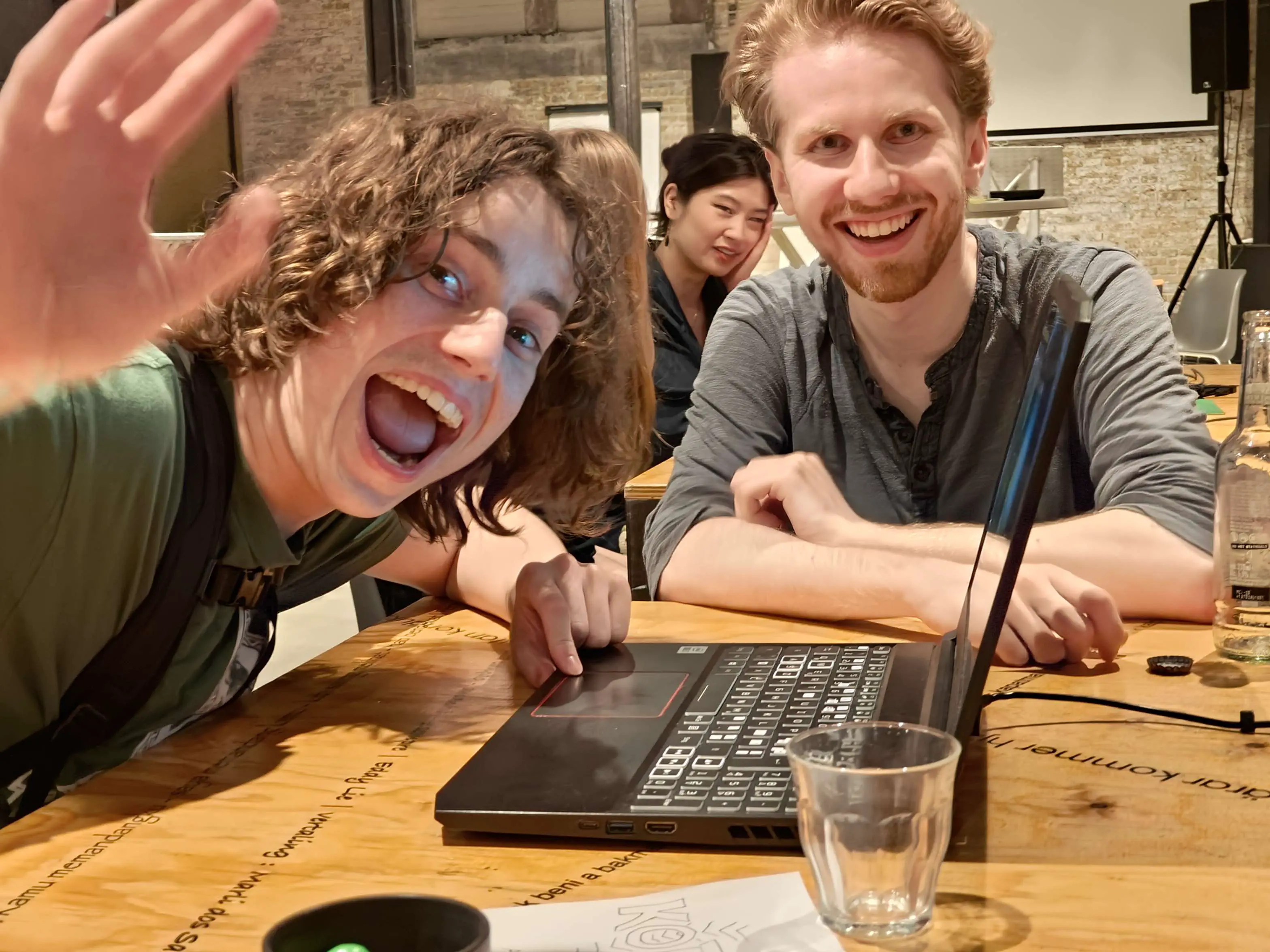 Photo taken during the Open Dev Mix, two people are waving and smiling for the camera behind a laptop